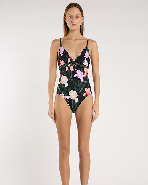 A person standing against a light neutral background, showing a black floral one piece swimsuit with thin spaghetti straps, a V-neckline, and an underbust seam. The fabric features pink and lavender floral prints with green stems and leaves.