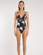 A person standing against a light neutral background, showing a black floral one piece swimsuit with thin spaghetti straps, a V-neckline, and an underbust seam. The fabric features pink and lavender floral prints with green stems and leaves.