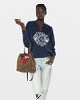 A person wearing a navy blue sweatshirt with a large white graphic reading "VOLTAIRE PARIS" along with a circular emblem and palm tree illustrations. The sweatshirt has a split crew neckline and relaxed fit. The person is also wearing light-wash distressed jeans and holding a brown leather handbag with a red and white charm attached. The background is a plain light grey.