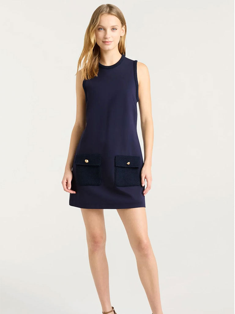 A woman wearing a sleeveless, navy shift dress that falls above the knee. The dress features a simple round neckline and two large textured front pockets with button details near the hem. She stands against a neutral background with her arms relaxed at her sides.