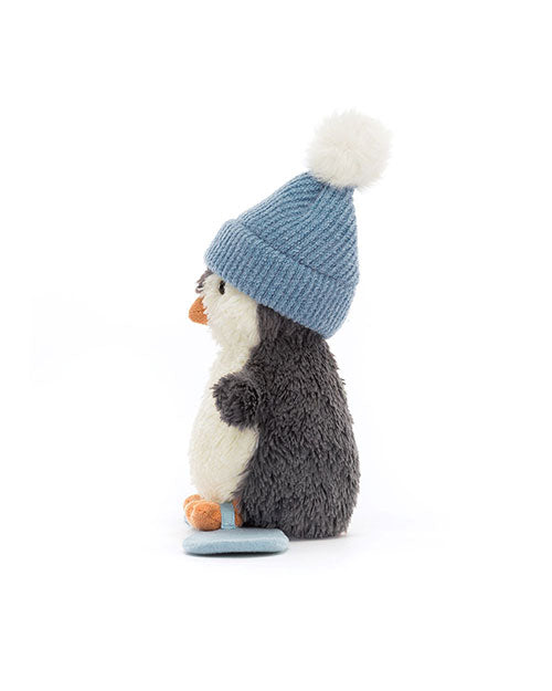 Plush toy penguin with a white belly, black back, orange beak and feet, wearing a blue knit hat with a white pom-pom, shown in profile view facing left.