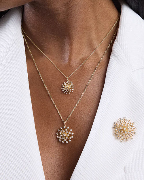 Close-up of layered gold necklaces featuring two starburst pendants adorned with small diamonds, paired with a matching gold starburst brooch pinned to a white textured blazer.