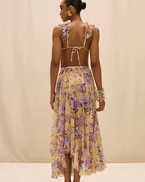 Back view of a person wearing a Flowy midi skirt in a warm yellow tone, featuring a soft floral print in shades of purple and lavender. The skirt has a high waistband and an asymmetrical hem with a ruffled edge, photographed against a neutral background in a mid-length view.
