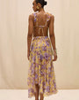 Back view of a person wearing a Flowy midi skirt in a warm yellow tone, featuring a soft floral print in shades of purple and lavender. The skirt has a high waistband and an asymmetrical hem with a ruffled edge, photographed against a neutral background in a mid-length view.