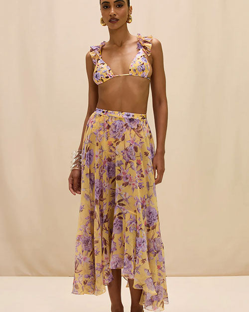 person wearing a flowy midi skirt in a warm yellow tone, featuring a soft floral print in shades of purple and lavender. The skirt has a high waistband and an asymmetrical hem with a ruffled edge, photographed against a neutral background in a mid-length view.