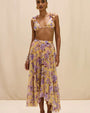 person wearing a flowy midi skirt in a warm yellow tone, featuring a soft floral print in shades of purple and lavender. The skirt has a high waistband and an asymmetrical hem with a ruffled edge, photographed against a neutral background in a mid-length view.