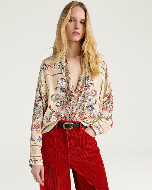 Waist-up studio image of a person wearing a cream-colored blouse with an intricate paisley print in shades of blue, red, and orange. The long-sleeve blouse features a relaxed fit, a ruffled stand collar, and a partially open neckline with gathered detailing at the front. The blouse is tucked into high-waisted red corduroy pants with a black belt and gold-tone buckle. Displayed against a light gray background.