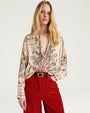 Waist-up studio image of a person wearing a cream-colored blouse with an intricate paisley print in shades of blue, red, and orange. The long-sleeve blouse features a relaxed fit, a ruffled stand collar, and a partially open neckline with gathered detailing at the front. The blouse is tucked into high-waisted red corduroy pants with a black belt and gold-tone buckle. Displayed against a light gray background.