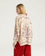 Back view of a cream-colored long-sleeve blouse featuring an allover paisley print in shades of blue, red, and orange. The blouse has a relaxed, slightly oversized fit with a pointed collar, dropped shoulders, and a curved hem. The ornate paisley motif is prominently displayed across the back and sleeves. Styled with high-waisted red corduroy pants against a light gray background.