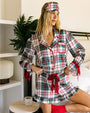 Person wearing a plaid pajama set with red ribbon accents, matching sleep mask worn as a headband, and red socks, posing beside a bed in a bright indoor bedroom setting.