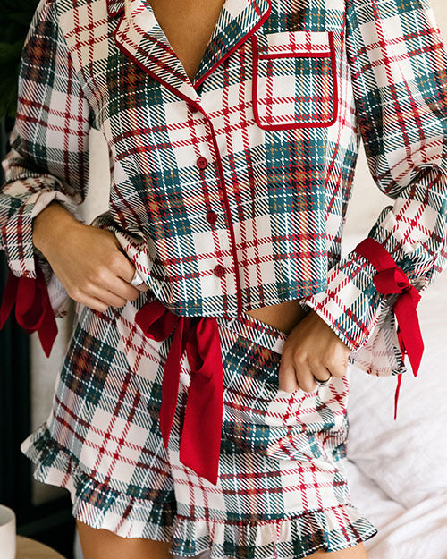 Close-up of a plaid pajama set featuring a button-front top with contrast piping, a chest pocket, red ribbon ties at the cuffs and waist, and a matching ruffled shorts skirt, shown in a bedroom setting.