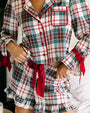Close-up of a plaid pajama set featuring a button-front top with contrast piping, a chest pocket, red ribbon ties at the cuffs and waist, and a matching ruffled shorts skirt, shown in a bedroom setting.