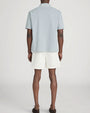 Rear-view studio image of a person standing against a light neutral background, wearing a light sage-green short-sleeve polo shirt with a soft collar and a relaxed fit, paired with off-white drawstring shorts. The shorts fall above the knee, and the outfit is styled with dark slip-on shoes, photographed in a full-length, back-facing pose.