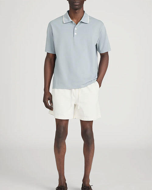 Person standing in a studio against a light neutral background, wearing a light sage-green short-sleeve polo shirt with a soft collar and button placket, paired with off-white drawstring shorts. One hand is placed in a pocket, and the image is photographed in a full-length, front-facing pose.