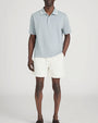 Person standing in a studio against a light neutral background, wearing a light sage-green short-sleeve polo shirt with a soft collar and button placket, paired with off-white drawstring shorts. One hand is placed in a pocket, and the image is photographed in a full-length, front-facing pose.
