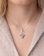 Close-up of two layered necklaces featuring animal-shaped pendants. The top necklace has a round pendant resembling a pomeranian's face, encrusted with white pavé-set gemstones and black gemstone eyes, on a gold chain. The bottom necklace features a dog’s head pendant with similar pavé detailing, on a silver chain.