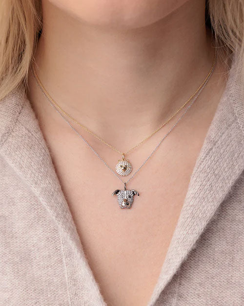 Close-up of two layered necklaces featuring animal-shaped pendants. The top necklace has a round pendant resembling a pomeranian's face, encrusted with white pavé-set gemstones and black gemstone eyes, on a gold chain. The bottom necklace features a dog’s head pendant with similar pavé detailing, on a silver chain.