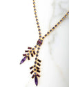 Close-up of a gold-toned necklace with a Y-shaped design, featuring purple baguette-cut crystals and marquise-shaped stones arranged in a branch-like pattern, displayed on a white marble surface.