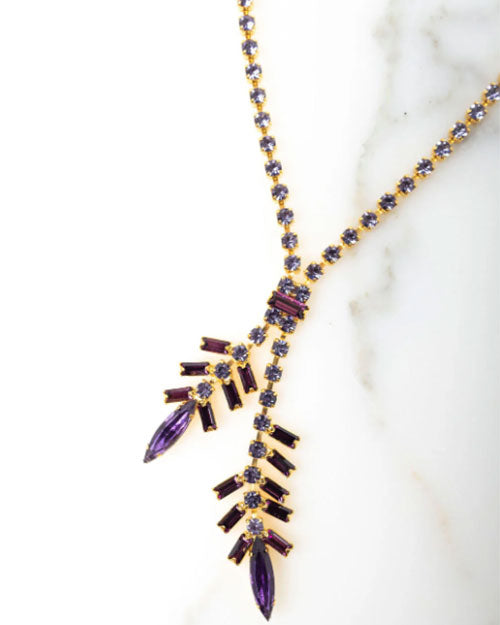 Close-up of a gold-toned necklace with a Y-shaped design, featuring purple baguette-cut crystals and marquise-shaped stones arranged in a branch-like pattern, displayed on a white marble surface.