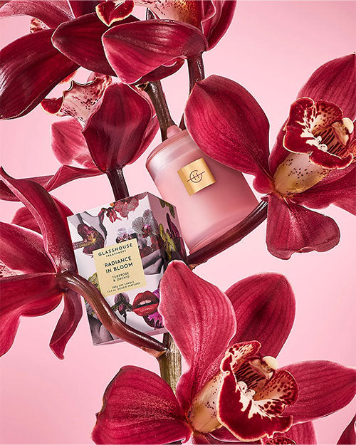 Pink glass candle and matching product box nestled among deep red orchid flowers against a soft pink background, featuring floral artwork and branding on the packaging.