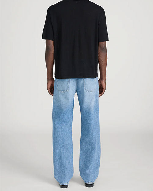 Back view of a person standing in a studio against a light neutral background, wearing a black short-sleeve polo shirt with a soft collar and relaxed fit, paired with light-wash straight-leg jeans. The outfit is styled with dark shoes, and the image is photographed in a full-length, front-facing pose.