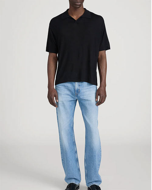 Person standing in a studio against a light neutral background, wearing a black short-sleeve polo shirt with a soft collar and relaxed fit, paired with light-wash straight-leg jeans. The outfit is styled with dark shoes, and the image is photographed in a full-length, front-facing pose.