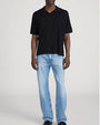 Person standing in a studio against a light neutral background, wearing a black short-sleeve polo shirt with a soft collar and relaxed fit, paired with light-wash straight-leg jeans. The outfit is styled with dark shoes, and the image is photographed in a full-length, front-facing pose.