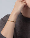 Close-up of a wrist wearing a sleek, polished gold bangle bracelet with a minimalist design, paired with a dark gray knit top.