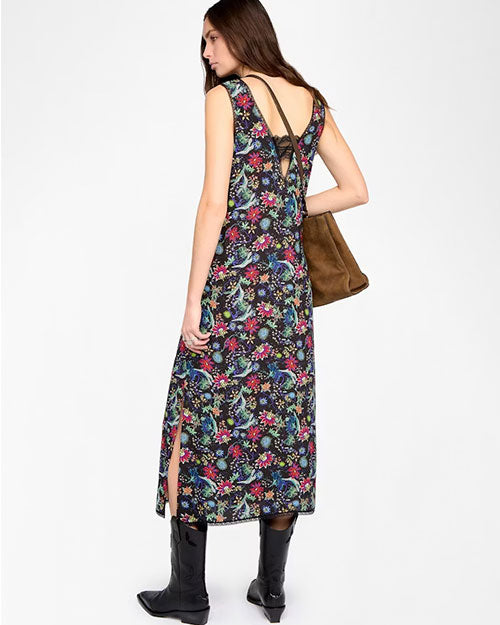 A person is standing against a plain white background wearing a sleeveless, ankle-length black dress with a colorful floral and botanical print in shades of green, pink, blue, and yellow. The dress has a deep V-back design with a crisscross strap detail and a side slit on the left. The outfit is paired with black pointed-toe cowboy boots and accessorized with a large brown handbag carried on the left shoulder.