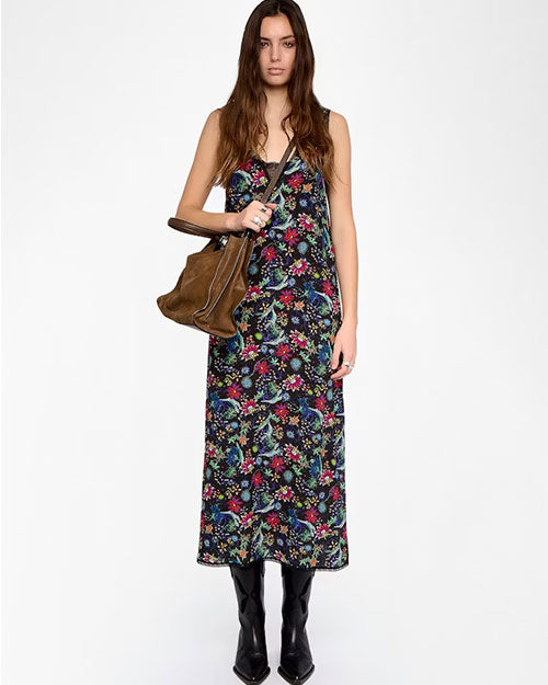 A person is standing against a plain white background wearing a sleeveless, ankle-length black dress with a colorful floral and botanical print featuring shades of green, pink, blue, and yellow. The outfit is paired with shiny black knee-high boots and accessorized with a large brown handbag held on the left arm. The hair is long and wavy, falling over the shoulders.