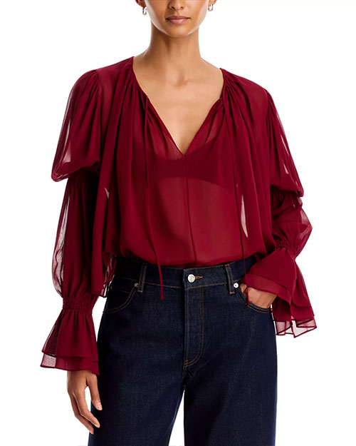 Sheer burgundy blouse with a V-neckline, gathered shoulders, voluminous long sleeves, and ruffled cuffs, tucked into dark-wash jeans against a light gray background.