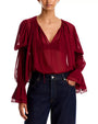 Sheer burgundy blouse with a V-neckline, gathered shoulders, voluminous long sleeves, and ruffled cuffs, tucked into dark-wash jeans against a light gray background.