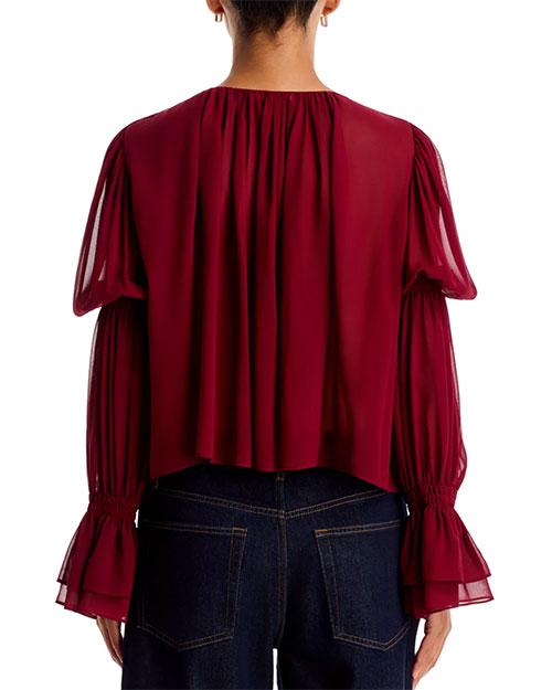 Sheer burgundy blouse with a V-neckline, gathered shoulders, voluminous long sleeves, and ruffled cuffs, tucked into dark-wash jeans against a light gray background.