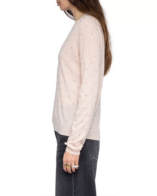  A person is shown from the side wearing a light pink long-sleeve sweater with small, evenly spaced rhinestone embellishments throughout the fabric. The sweater has a relaxed fit and is paired with high-waisted, faded black jeans. The left hand is visible, adorned with multiple gold rings, and the hair is long and straight, falling over the shoulder.