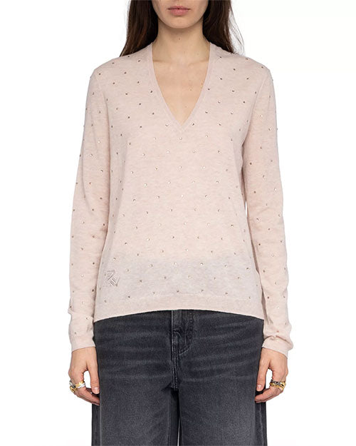  A person is standing against a plain white background wearing a light pink long-sleeve sweater with a V-neckline. The sweater is embellished with small, evenly spaced rhinestones throughout the fabric, adding a subtle sparkle. The outfit is paired with high-waisted, faded black jeans. Multiple rings are visible on both hands, which are relaxed by the sides.
