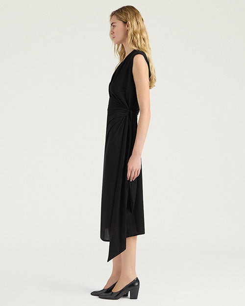 Side view of a black sleeveless midi dress with a draped V-neckline and gathered wrap-effect detailing at the waist. The dress features a slim silhouette and an asymmetrical overlay that extends below the hem. Styled with black pumps and shown against a light gray background.