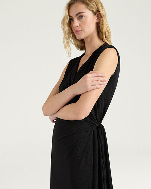 Close-up view of a black sleeveless dress featuring a draped V-neckline and gathered wrap-effect detailing at the side waist. The dress has a soft, fluid silhouette with subtle ruching across the front and a tie detail extending from the waist. Shown against a light gray background.