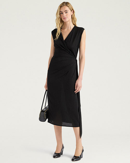Front view of a black sleeveless midi dress with a draped V-neckline and gathered wrap-effect detailing at the side waist. The dress features a slim silhouette, asymmetrical hem, and side slit. Styled with black pumps and a black handbag, and shown against a light gray background.
