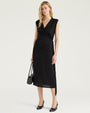 Front view of a black sleeveless midi dress with a draped V-neckline and gathered wrap-effect detailing at the side waist. The dress features a slim silhouette, asymmetrical hem, and side slit. Styled with black pumps and a black handbag, and shown against a light gray background.
