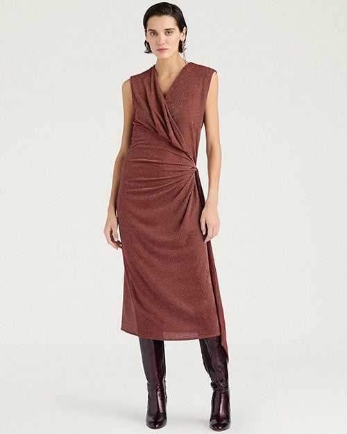 Front view of a sleeveless burgundy midi dress with a subtle metallic shimmer. The dress features a draped V-neckline, gathered wrap-effect detailing at the waist, and an asymmetrical hem with a side slit. Styled with knee-high burgundy boots and shown against a light gray background.