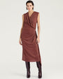 Front view of a sleeveless burgundy midi dress with a subtle metallic shimmer. The dress features a draped V-neckline, gathered wrap-effect detailing at the waist, and an asymmetrical hem with a side slit. Styled with knee-high burgundy boots and shown against a light gray background.