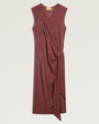 Product image of a sleeveless burgundy midi dress with a subtle metallic shimmer. The dress features a draped V-neckline, wrap-effect construction, and gathered tie detail at the side waist that creates soft pleating through the skirt. An asymmetrical overlay extends from the waist to the hem. Shown laid flat against a light gray background.