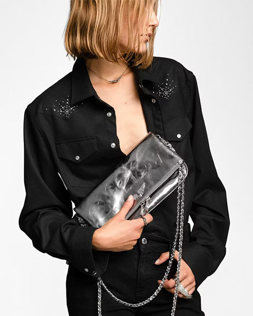 Person wearing a black button-up shirt with pointed collar and flap chest pockets, featuring small stud embellishments near the shoulders. The shirt is styled partially open and paired with black bottoms. The person is holding a metallic silver clutch bag with a shiny, textured finish and a silver chain strap