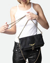 Person wearing a white ribbed tank top and black textured pants, holding a black leather clutch bag with a gold wing emblem and a gold chain strap.