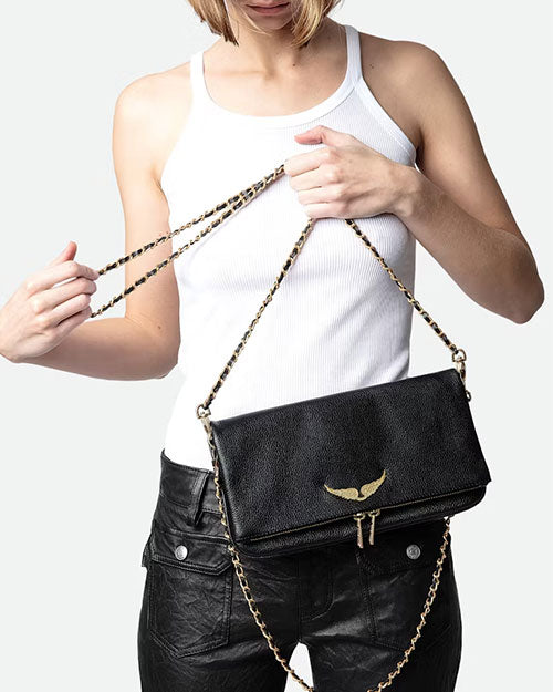 Person wearing a white ribbed tank top and black textured pants, holding a black leather clutch bag with a gold wing emblem and a gold chain strap.