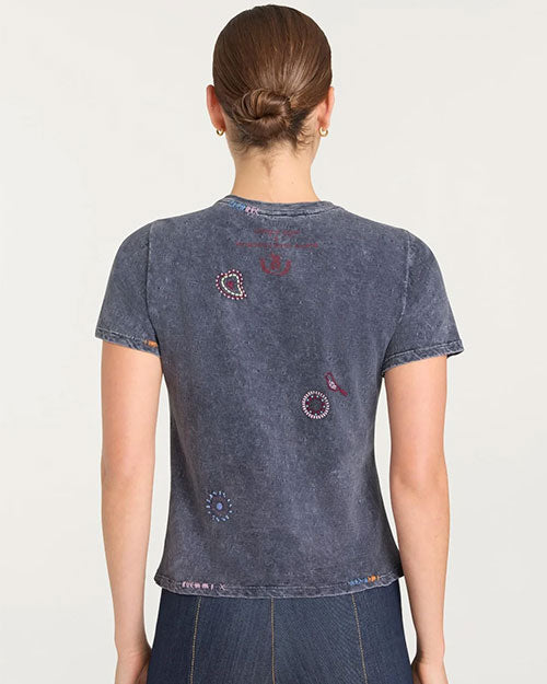Back view of a faded black short-sleeve graphic T-shirt featuring scattered embroidered designs in red, white, and purple tones, including paisley shapes, circular patterns, and a horseshoe motif. The shirt has a slightly distressed texture and is paired with dark denim bottoms.