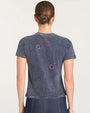 Back view of a faded black short-sleeve graphic T-shirt featuring scattered embroidered designs in red, white, and purple tones, including paisley shapes, circular patterns, and a horseshoe motif. The shirt has a slightly distressed texture and is paired with dark denim bottoms.