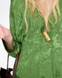  Close-up of a green textured dress with a subtle abstract pattern. The dress has a V-neckline with a small tie detail at the center. A gold pendant necklace with a rectangular, hammered design hangs down the front. Part of a brown leather handbag strap is visible on the left side.