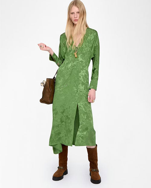 A person is standing against a plain white background wearing a long green textured dress with a front slit. The outfit is accessorized with a gold pendant necklace, a brown handbag held on the left side, and brown knee-high suede boots with buckles. The dress has long sleeves and an asymmetrical hemline.