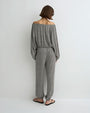 Back view of a person modeling a long-sleeve off-the-shoulder jumpsuit with a small-scale black and white pattern, featuring a relaxed fit, elastic waistband, and loose straight-leg pants, paired with black flat sandals against a plain light background.